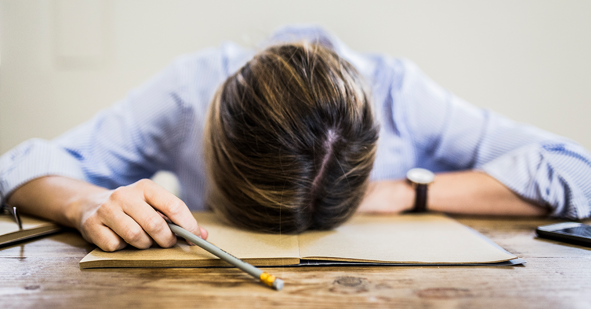 Person with head down on desk holding a pencil